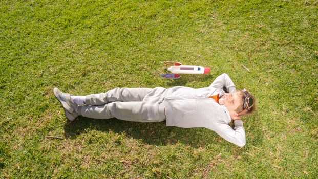 Young boy in a gray sweater daydreaming about space travel, lying on grass with toy rocket.