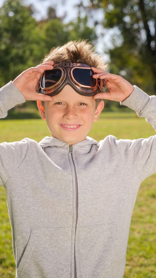 Free Smiling boy in a gray jacket with vintage aviator goggles in a sunny park setting. Stock Photo