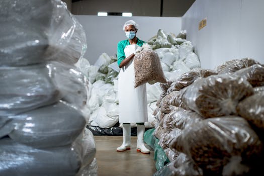 A worker in a face mask and apron organizes stacks of industrial plastic bags in a warehouse.