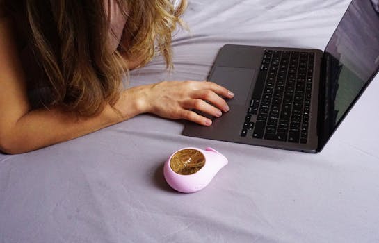 A woman lies on her bed using a laptop with a small device nearby, depicting a casual work-from-home setup.