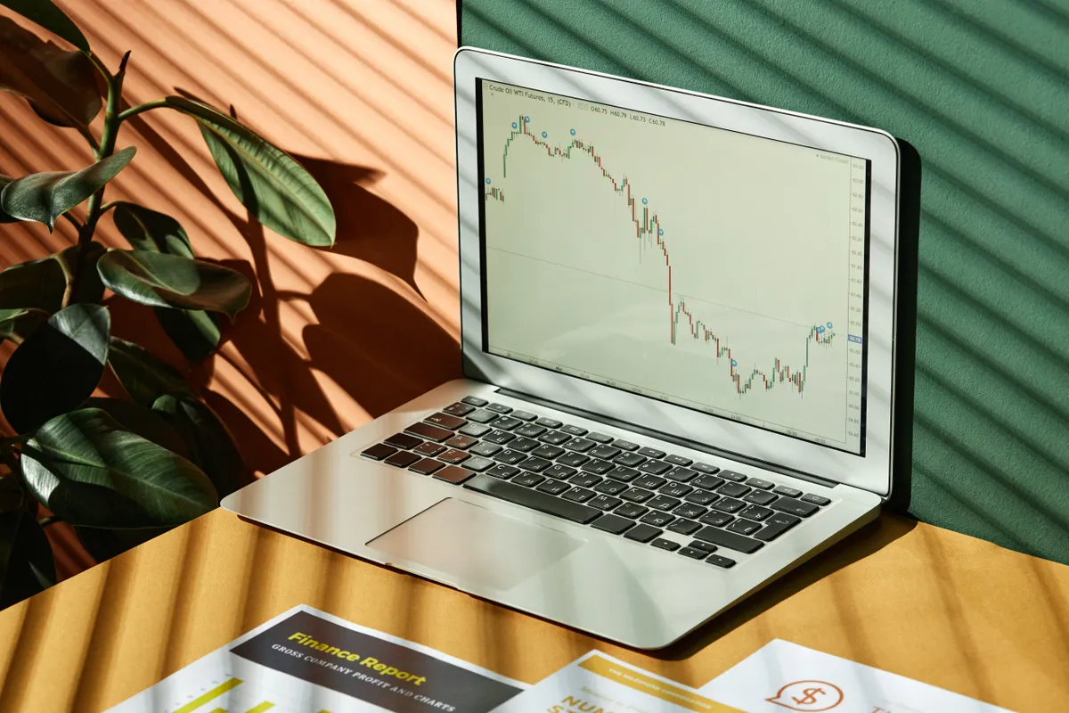 A silver laptop on a desk in striped sunlight shows a falling price chart, with finance printouts in front of it.