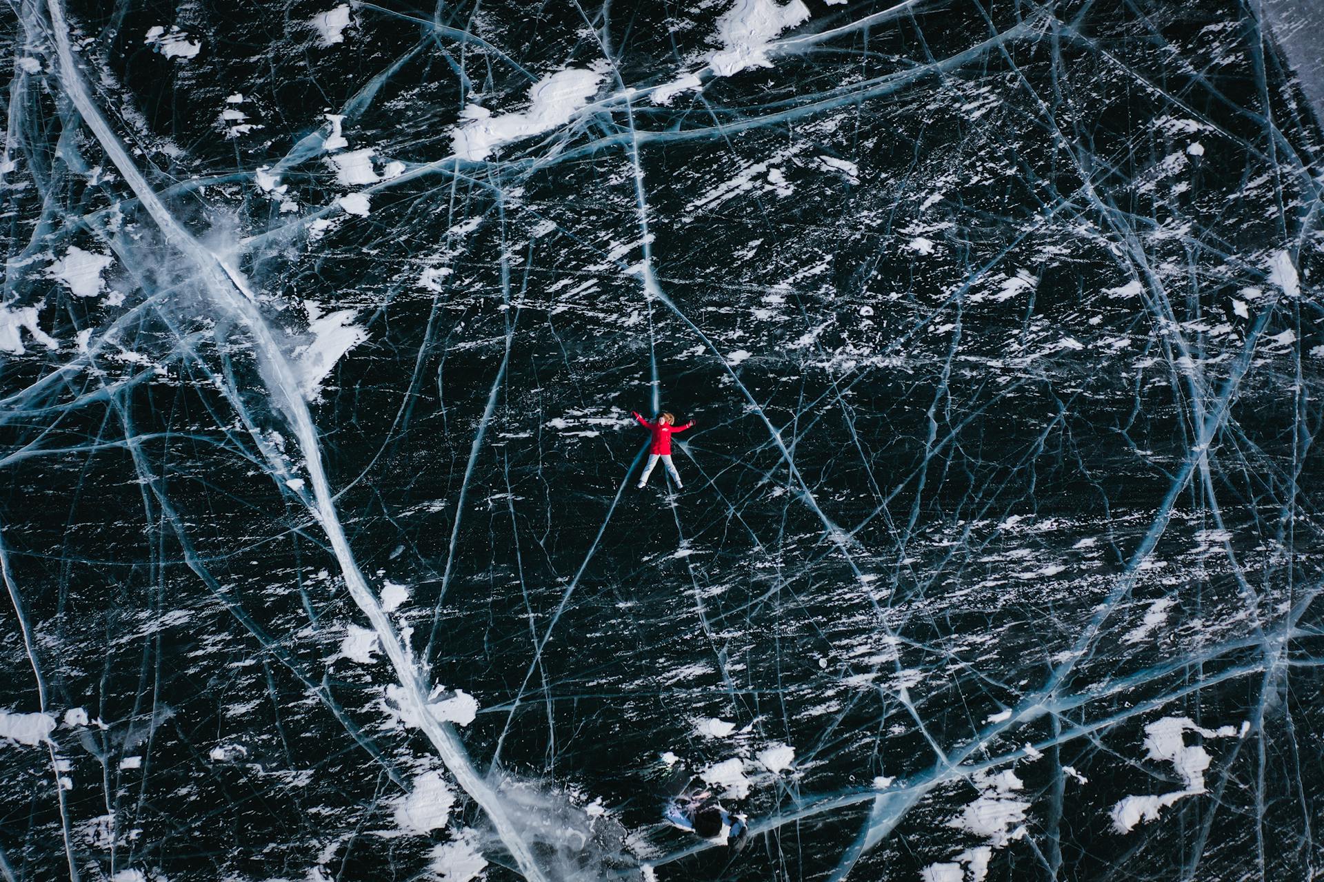 Drone view of anonymous tourist lying on frozen river with cracks during winter trip in daytime