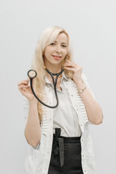 Blonde female doctor smiling while holding a stethoscope, wearing a white top on a plain background.