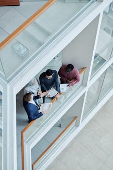 High-angle view of diverse colleagues discussing documents in a modern, glass-structured office.
