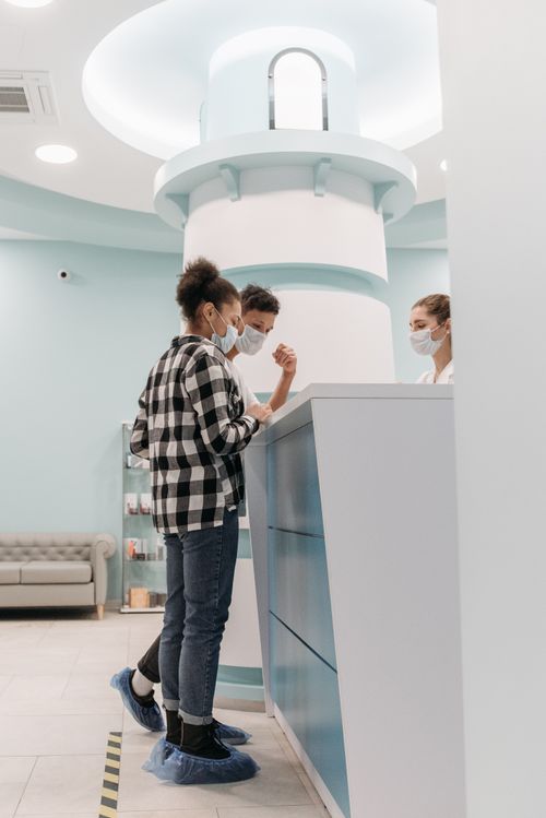 Free Two people in a clinic reception discussing with a medical assistant, all wearing masks and shoe covers. Stock Photo
