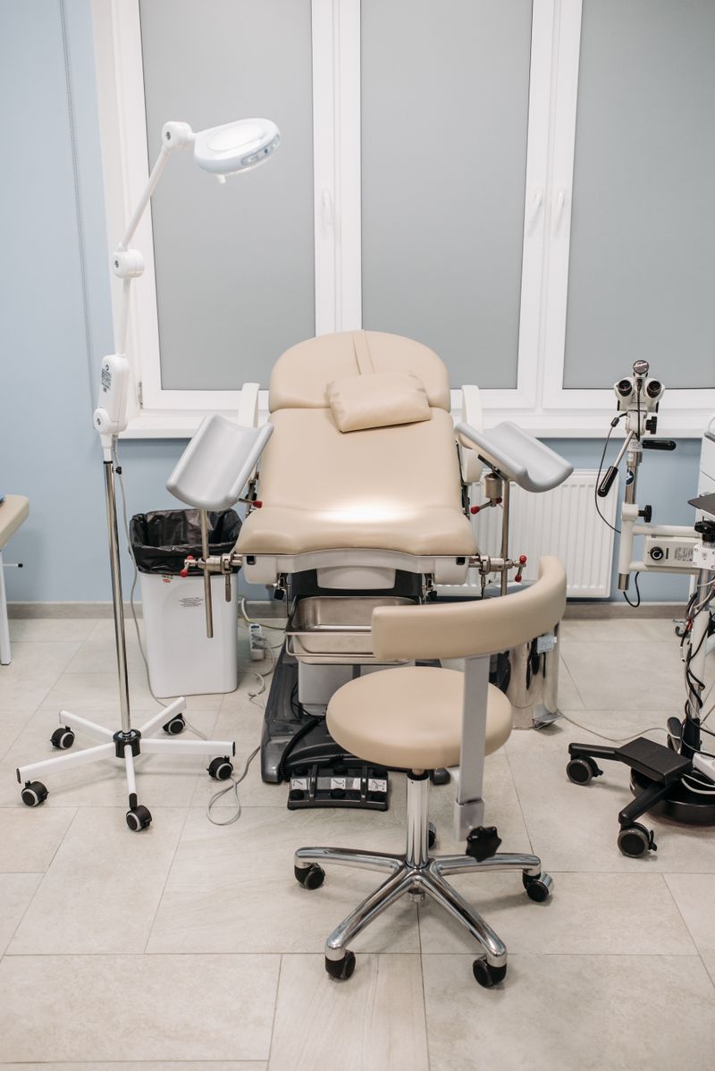 An empty gynecological examination room with a tan padded exam chair with leg supports, a swing-arm exam lamp, and ancillary medical equipment against pale blue walls and tile floor.