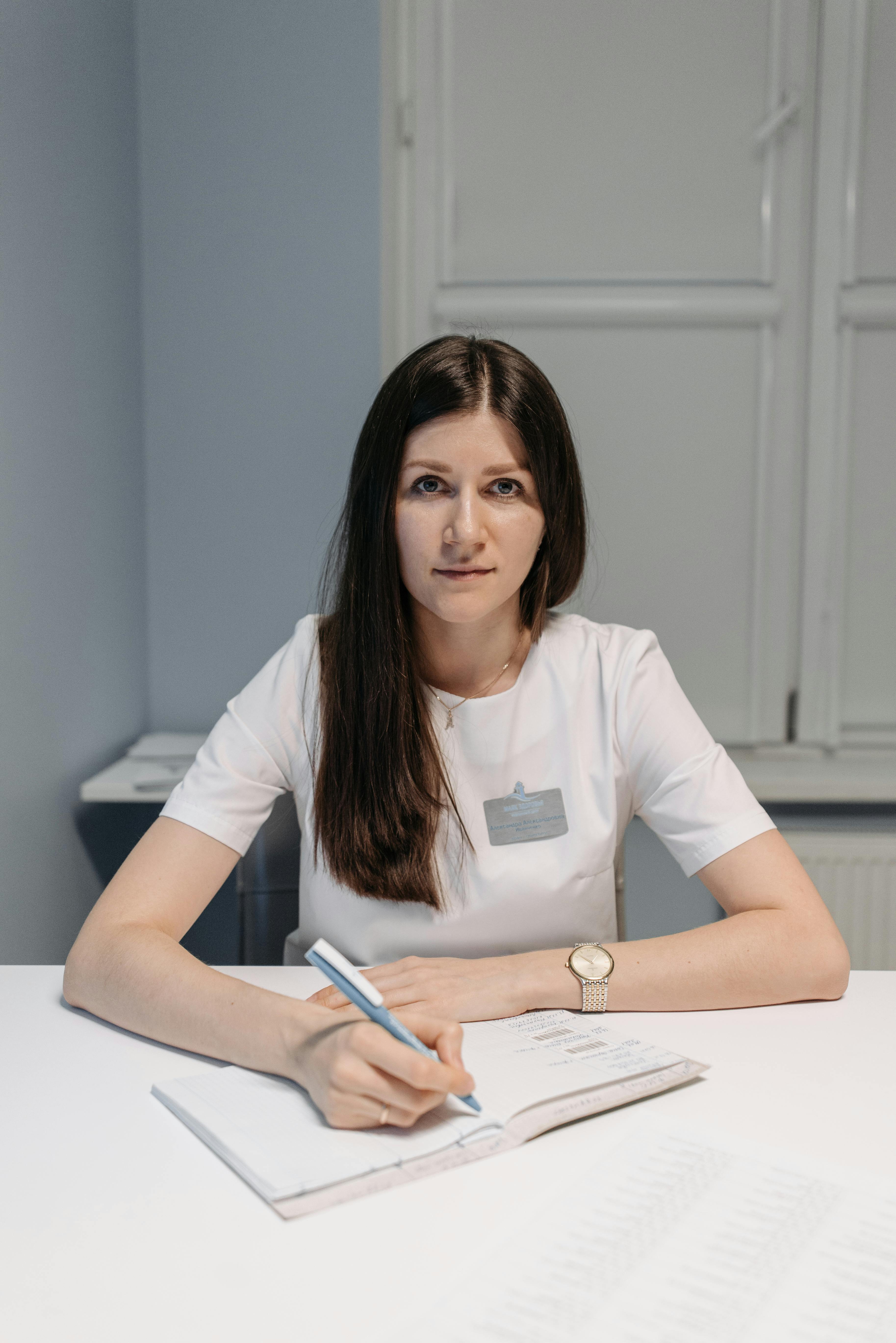 A woman in a white uniform writing in a notebook at an indoor office setting.