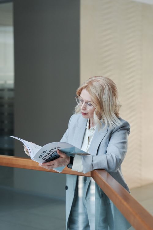 Free Adult woman in a gray blazer reading a book in a modern office setting. Stock Photo