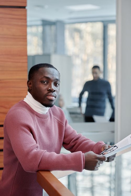 Free African American man dressed in a sweater holding a document in a stylish office setting. Stock Photo