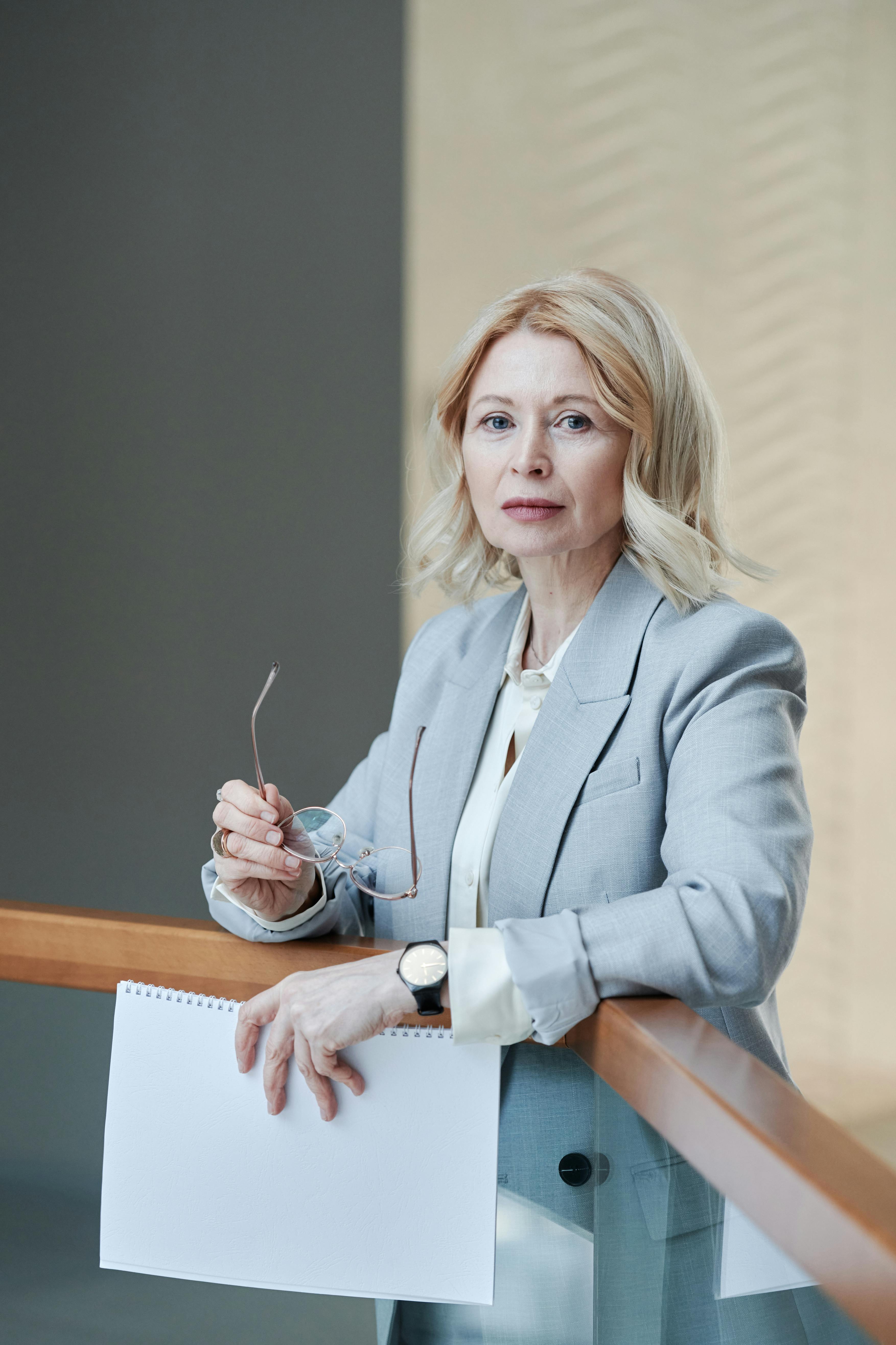 Senior caucasian woman in gray blazer holds papers in a modern workplace setting.