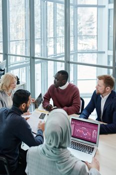 A diverse business team discusses reports and statistics around a wooden table with laptops indoors.