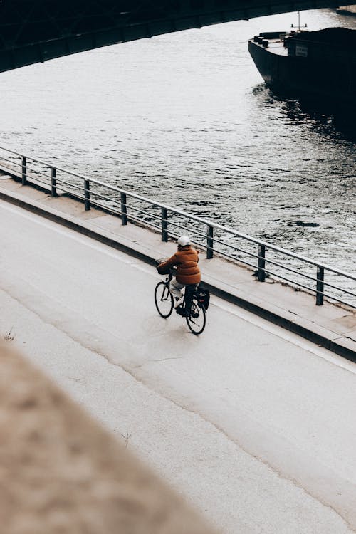 From above back view of anonymous bicyclist riding bike on fenced bridge above lake with ship