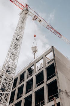 From below of modern hydraulic luffing jib tower crane working at construction site near modern unfinished multistory building under cloudy sky in city
