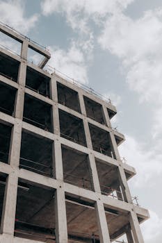 A modern multistory concrete building under construction, framed against a blue sky and clouds.