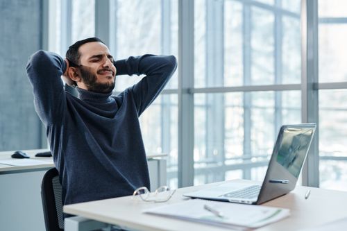 Free Man leaning back in office chair, appearing relaxed amidst a busy workday, reflecting stress management. Stock Photo
