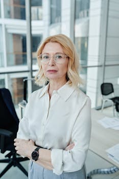 Professional woman with glasses standing confidently in a modern glass office environment.