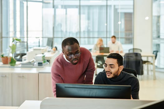 Two male colleagues working together at a computer in a modern office environment, discussing and collaborating on a project.