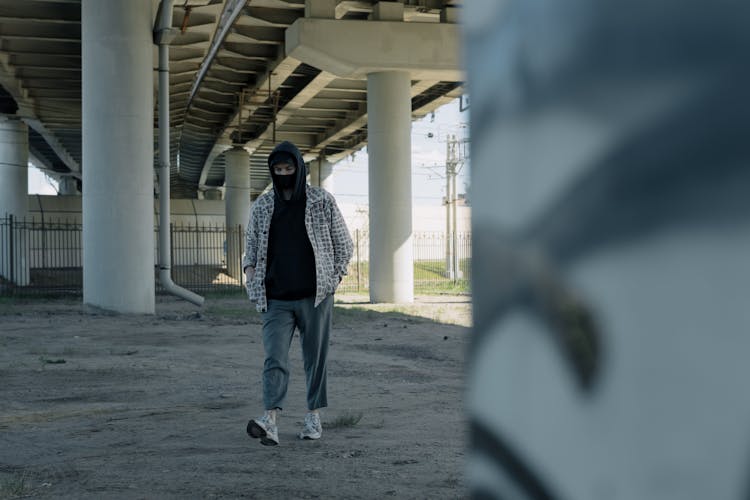 A Man Wearing Hoodie And Face Mask Walking Under A Bridge