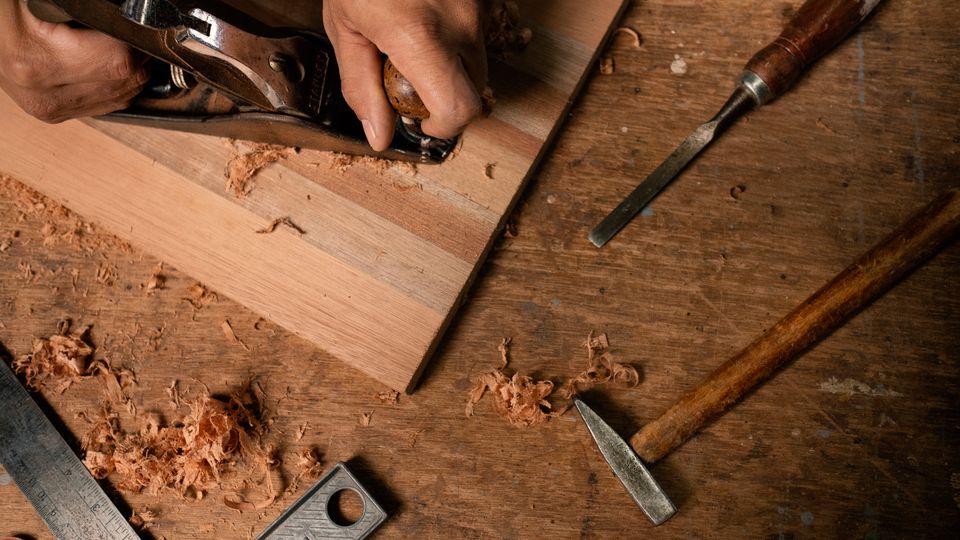 Hænder høvler en træplade på en snedkerbænk med mejsel, hammer og høvlspåner