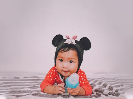 Cute baby wearing a mouse-themed hat, holding a bottle, and lying indoors on a striped blanket.