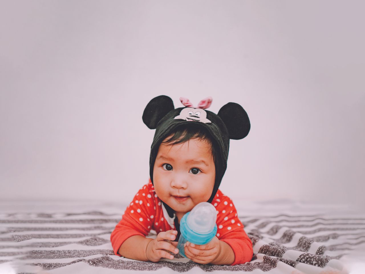 cute baby wearing a mouse themed hat holding a bottle and lying indoor