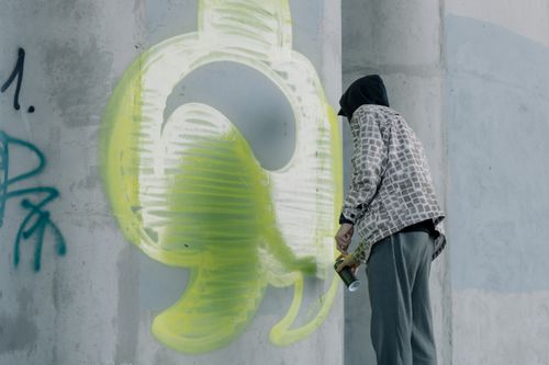 Free A hooded artist spray-paints vibrant graffiti on a concrete wall in an urban setting. Stock Photo