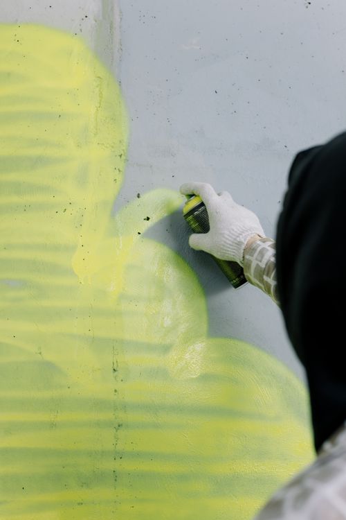 Free A person in gloves sprays yellow graffiti on a concrete wall, showcasing urban art. Stock Photo