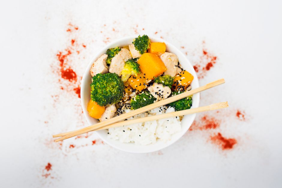A white bowl of steamed rice topped with glazed chicken pieces, broccoli florets, and julienned carrots, garnished with sliced green onions and sesame seeds.
