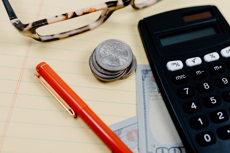 Close-up of a workspace with a calculator, coins, and glasses on a notepad.