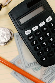Black calculator, coins, and cash on a yellow notepad with a pencil.