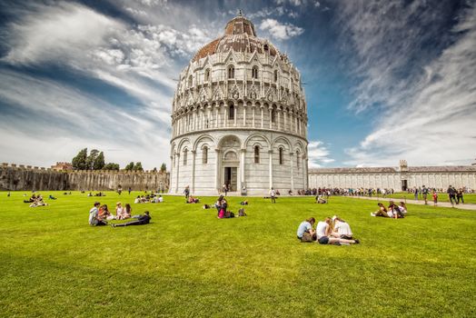 A vibrant scene at Pisa Baptistery with people enjoying the lush green surroundings under a bright sky.