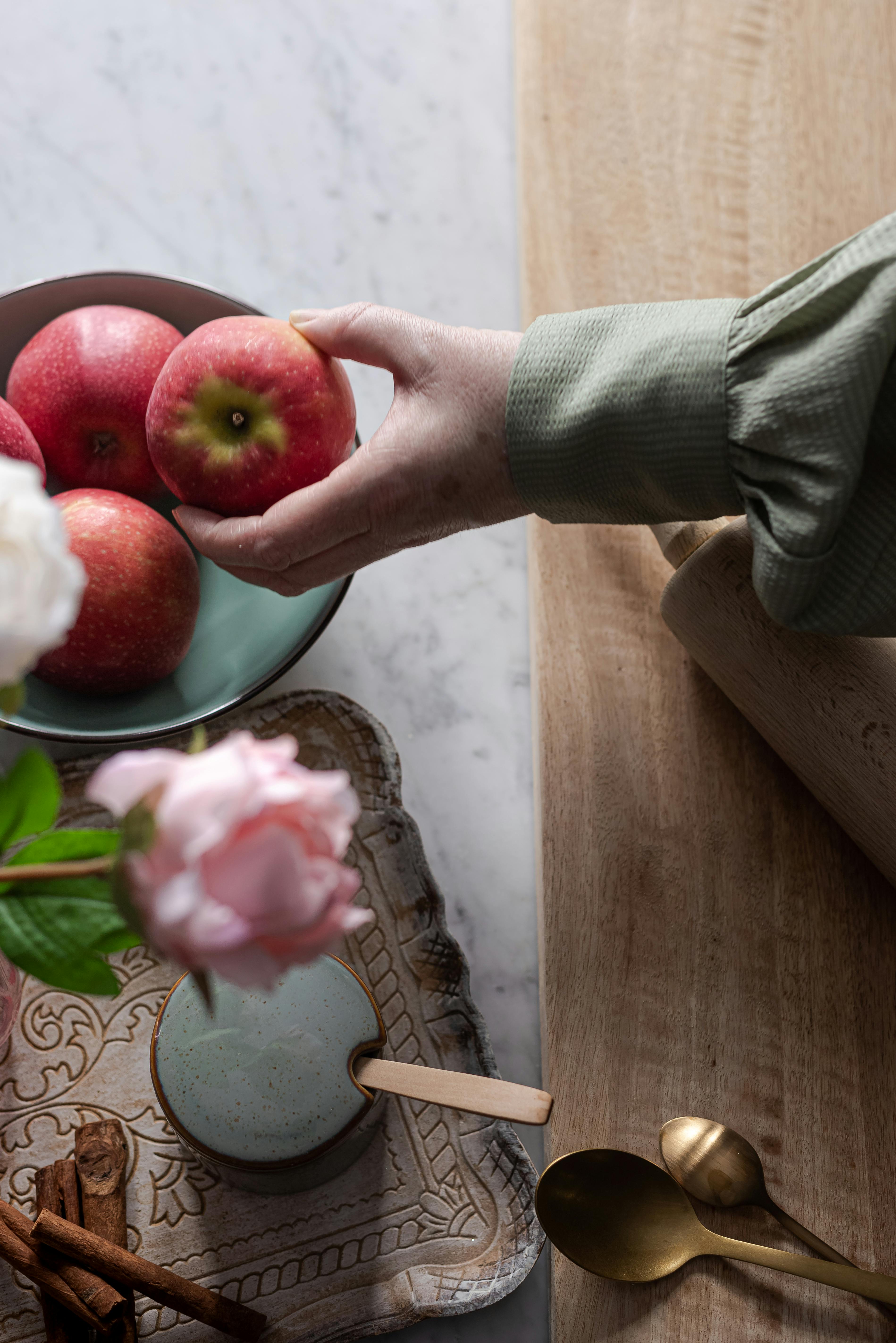 Top view of hand holding apple above wooden tray with flowers and kitchen utensils, artistic composition.