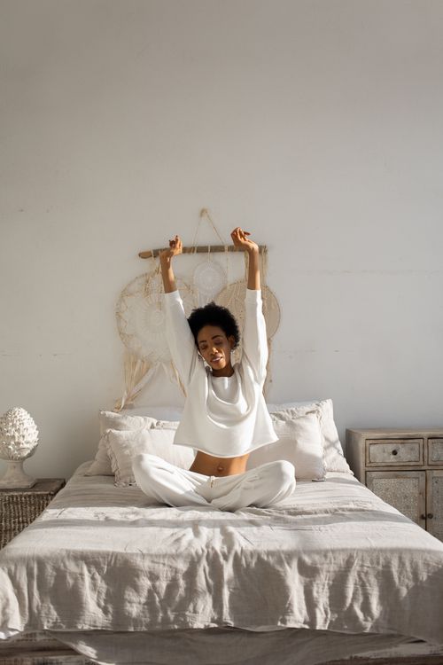 Free African American woman stretches in a sunlit, minimalist bedroom with neutral decor. Stock Photo