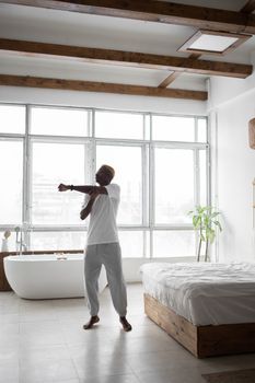 Man stretching his arm in a bright, minimalist bedroom with a bath tub and large windows.