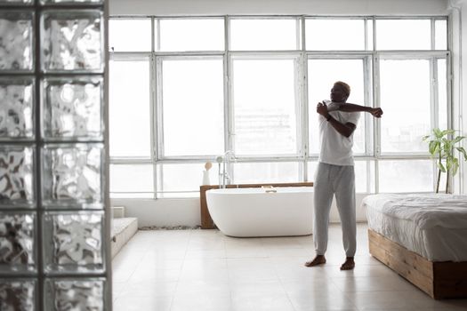 Adult man in white shirt stretches by a bathtub in a bright modern bedroom with glass windows.