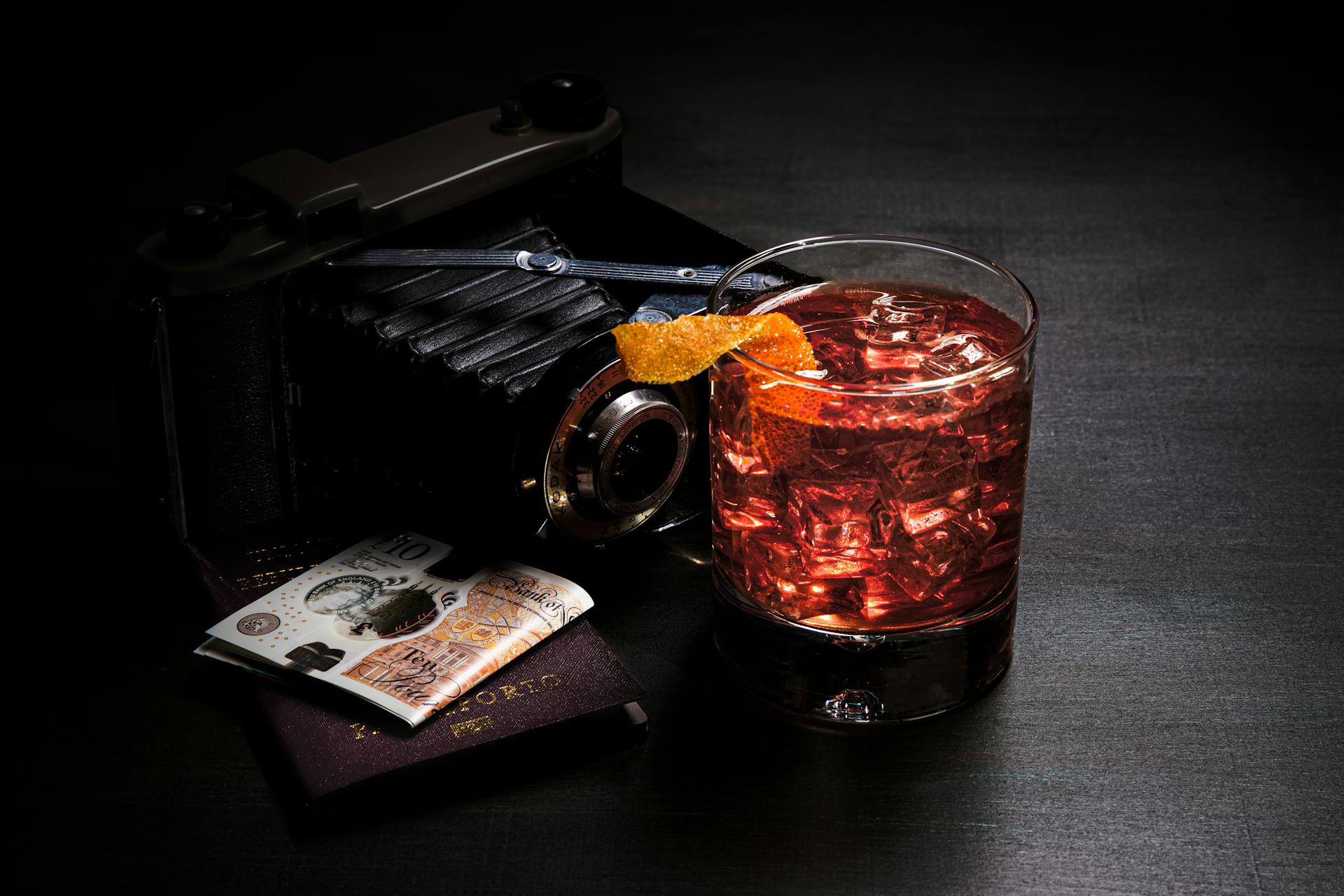 A vintage camera, cocktail with ice and cash create a moody still life on a dark background.