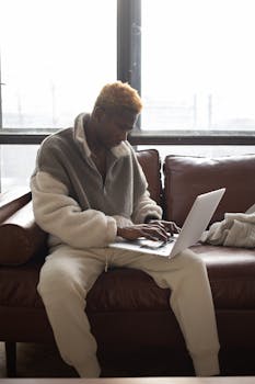 Adult man sitting on a leather couch using a laptop by the window, casual indoor setting.