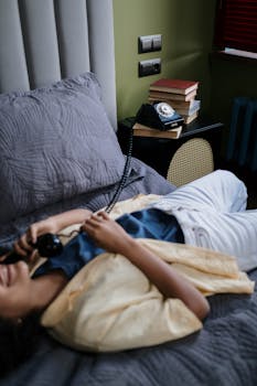 A relaxed woman lies on a bed, comfortably holding a vintage rotary telephone in a cozy indoor setting.