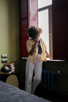 Woman with curly hair making a phone call by a window in a cozy room.