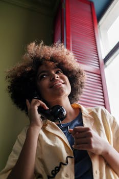 A young woman with afro hair talks on a vintage rotary phone near a window. Indoor shot.