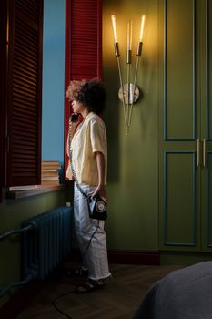Woman with afro hair standing by a window holding a vintage rotary phone in a stylish room.