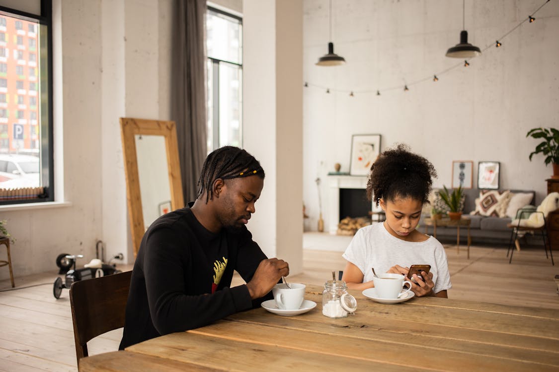Free Serious African American father having coffee at table with girl browsing mobile phone in flat Stock Photo