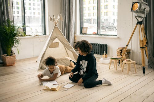 High angle of African American mother sitting on floor near girl reading book near play tent in studio