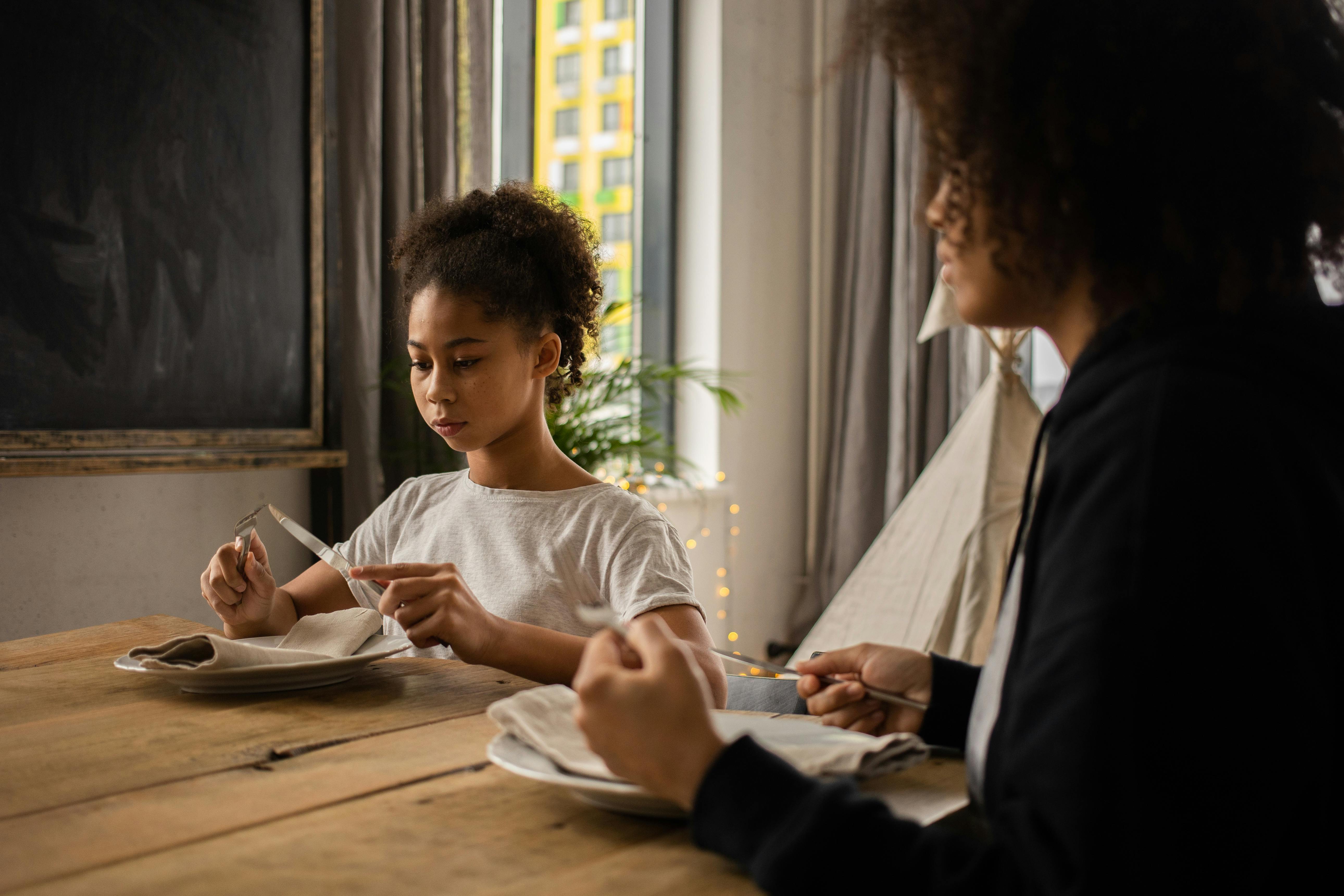 black girl at table with plate and cutlery near anonymous mother