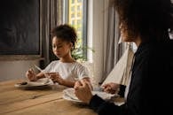 Black girl at table with plate and cutlery near anonymous mother