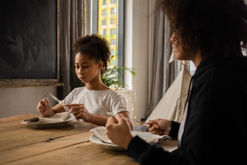 Free Serious African American girl with black faceless mother sitting at wooden table with plate and silverware while learning dining etiquette in room Stock Photo