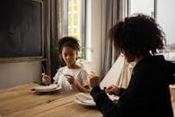 Pensive African American girl sitting at wooden table with plate and cutlery in hands while learning dining etiquette with anonymous mother