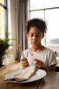 Serious African American girl sitting with knife and fork in hands near white plate with napkin while learning table manners