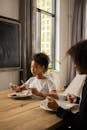 Serious African American girl with unrecognizable mother sitting at wooden table with plate and silverware in hands learning table manners