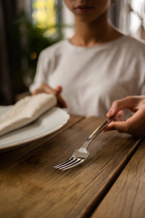 Crop unrecognizable female sitting at wooden table with white plate and fork while learning dining etiquette in room on blurred background